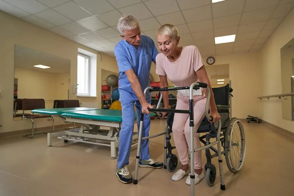 Photo of middle-aged woman holding walking frame while trying making steps with physiotherapist assistance in rehab center