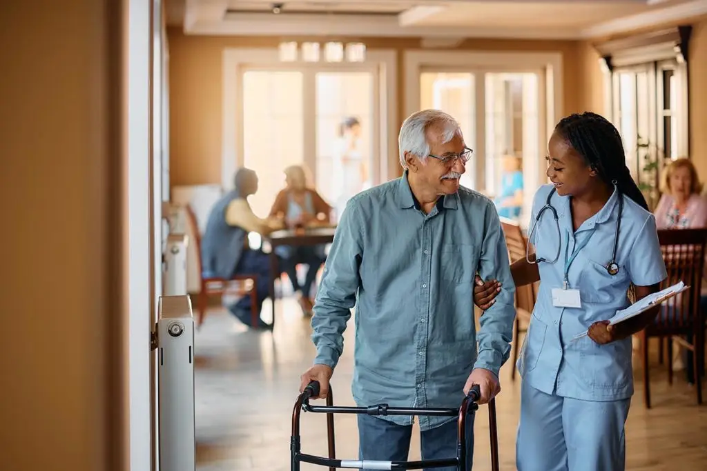 Happy senior man using mobility walker while female nurse is helping him in nursing home.