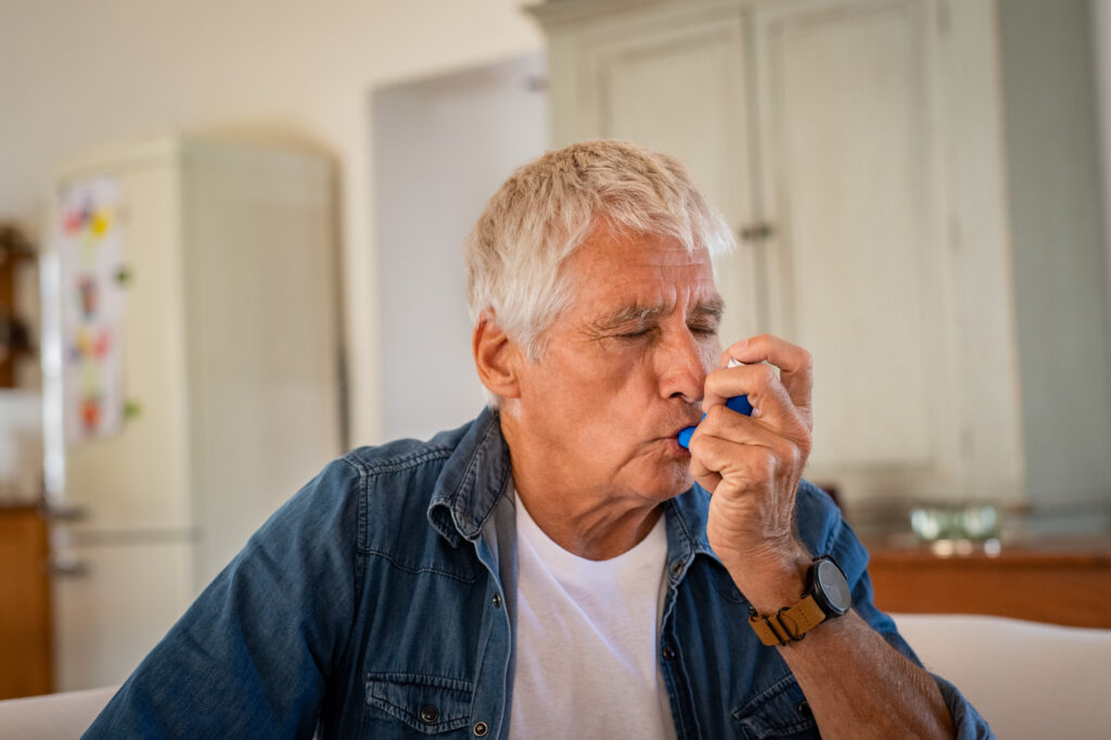 Senior man using asthma inhaler for relief an attack at home. Old man using asthma inhaler for preventing attack. Mature man using medical inhaler to prevent and treat wheezing and shortness of breath caused by allergy.
