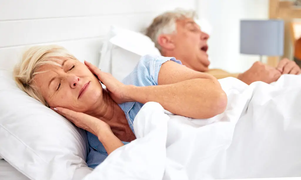 Senior woman in bed with hands covering her ears and eyes closed in frustration as her husband snores beside her.