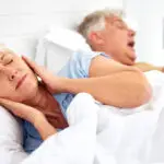 Senior woman in bed with hands covering her ears and eyes closed in frustration as her husband snores beside her.